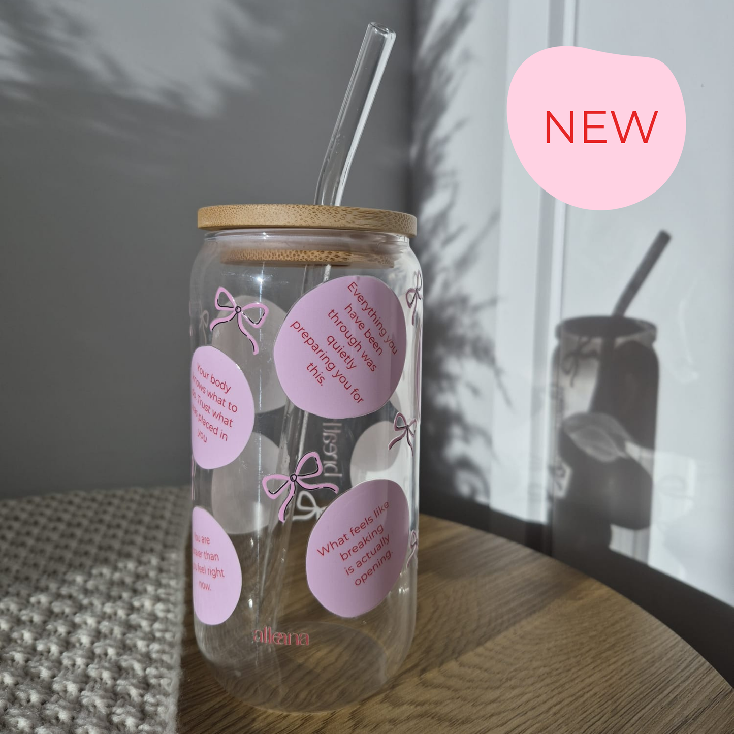 A clear glass can-shaped tumbler with a bamboo lid and clear straw, decorated with pink heart-shaped stickers and bow illustrations. It sits on a wooden table in a softly lit room, with a pink 'NEW' badge overlay in the top right.